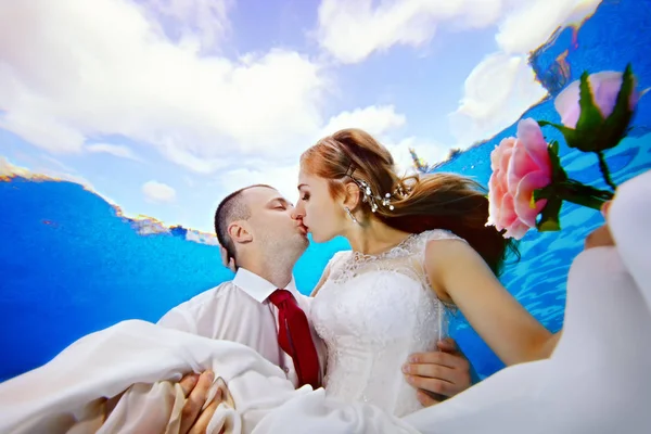 Очаровательная невеста с цветами в руке и жених целуется под водой на фоне облаков на поверхности. Портрет. Крупный план. Вид снизу. Подводная фотография. Горизонтальный вид — стоковое фото