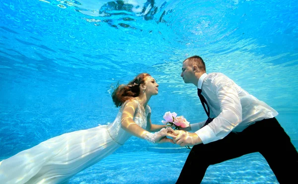 En vacker brud i en vit klänning simmar under vattnet till brudgummen och tar händerna. Pojken och flickan belyses av solen under vattnet. Porträtt. Koncept. Undervattensfotografering — Stockfoto