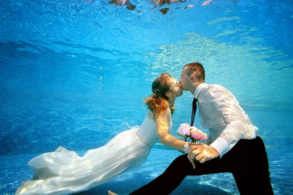 De mooie bruid en bruidegom houden handen en kus onderwater aan de onderkant van het zwembad op een zonnige dag. Portret. Concept. Onderwater bruiloft. Horizontale weergave — Stockfoto
