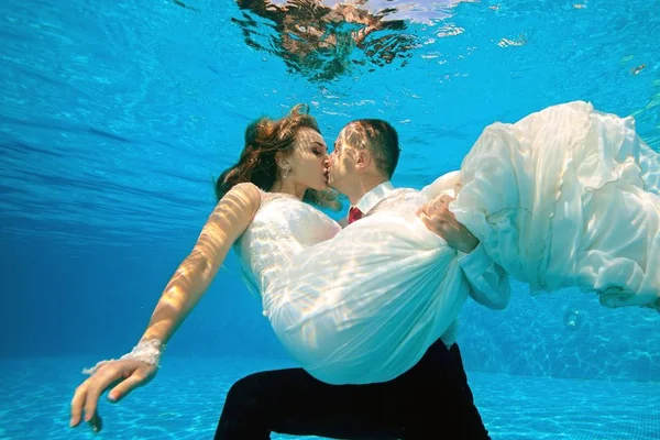 Lycklig brud och brudgum kysser under vattnet i poolen på en solig dag. Brudgummen kramar och håller bruden i armarna längst ner. Porträtt. Koncept. Bröllop under vattnet. Horisontell vy — Stockfoto