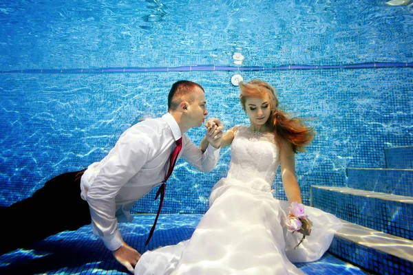 Ο γαμπρός φιλάει το χέρι της γοητευτικής νύφης υποβρύχια σε μια ηλιόλουστη μέρα. Κάθονται στον πάτο της πισίνας με νυφικά. Πορτρέτο. Γκρο πλαν. Έννοια. Γάμος κάτω από το νερό. Οριζόντια προβολή — Φωτογραφία Αρχείου