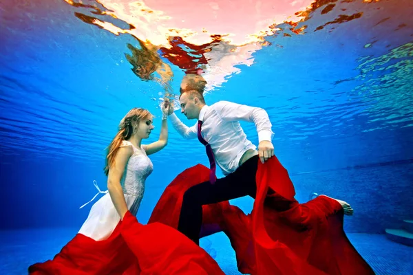 La hermosa novia y el novio bailan bajo el agua entre sí y posan con un paño rojo en sus manos sobre un fondo azul en un día soleado. Retrato. Concepto. Boda bajo el agua. Vista horizontal — Foto de Stock