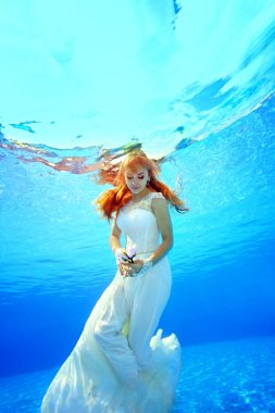 Güzel bir gelin yüzme ve güneşli bir günde beyaz bir elbise içinde havuzda sualtı poz. Çiçeği elinde tutuyor ve ona bakıyor. Portre. Sualtı fotoğrafçılığı