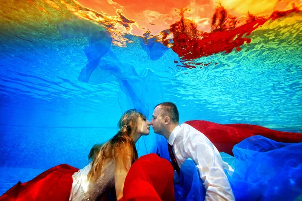 Fabulosa boda bajo el agua. Los novios nadan y se besan bajo el agua en la piscina al atardecer rodeados de tela roja y azul. Retrato. Concepto. Fotografía submarina. Vista horizontal — Foto de Stock