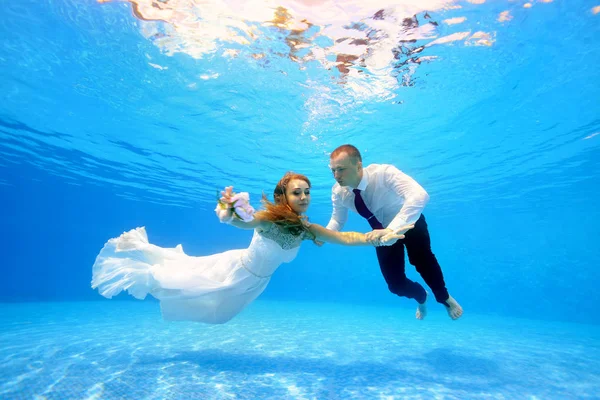 Lycklig brud och brudgum i vit klänning simma under vattnet i poolen. De flyter i det klara blå vattnet med sina armar utsträckta. Porträtt. Horisontell orientering — Stockfoto