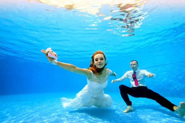 新娘穿着白色礼服和新郎游泳，在清澈的蓝色海水中的泳池底部用相机在水下摆姿势。肖像。水平方向 — 图库照片