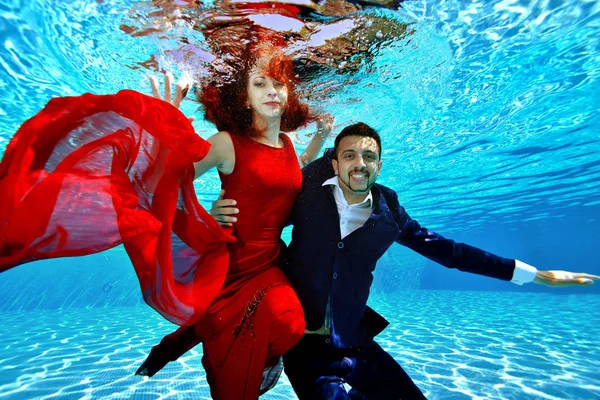 A guy and a girl in a red evening dress swim and play underwater in the pool on a Sunny day. They smile and pose for the camera on a blue background. Portrait. Fashionable underwater shooting