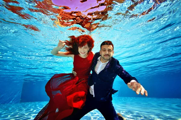 A beautiful girl in a red dress and a guy in a suit are swimming underwater in the pool on a Sunny day. They smile and pose for the camera on a blue background. Fashionable underwater shooting