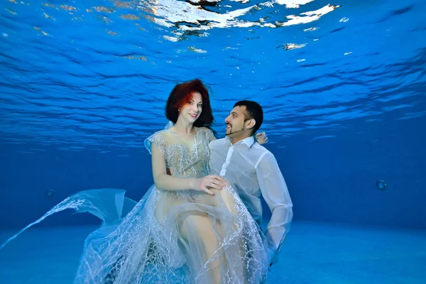 A cute girl in a transparent white dress sits under the water on the lap of an oriental guy and poses for the camera. Portrait. Conceptual photo. Wedding underwater — Stock Photo, Image