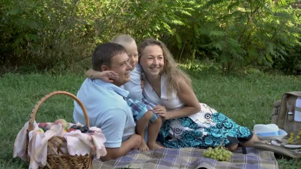 Un bambino allegro abbraccia suo padre e sua madre che sono seduti sull'erba del parco al tramonto e sorridono. Vacanza in famiglia nella natura. Ritratto. Concetto. 4K — Video Stock