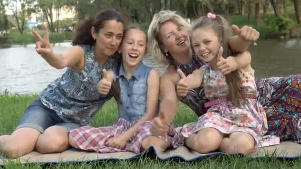 Крупним планом матері з молодими доньками грають і розважаються, сидячи на газоні в парку на заході сонця в літній день біля річки. Сімейний відпочинок на свіжому повітрі. Концепція сімейного щастя. 4-кілометровий — стокове відео
