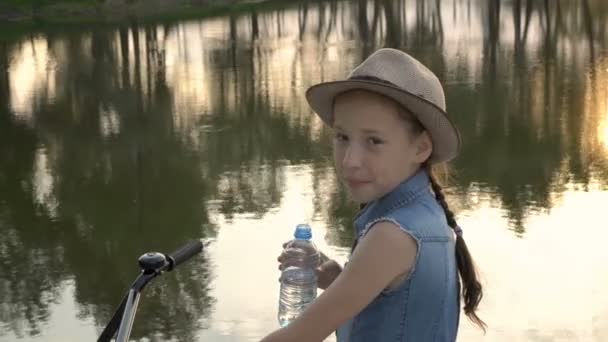 Linda niña sonriendo a la cámara y bebiendo agua al aire libre, en verano al atardecer en el fondo del río, sentada en una bicicleta. Retrato. Concepto. 4K — Vídeo de stock