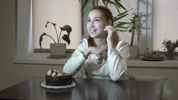 Menina pensativa sentada perto do bolo na mesa na cozinha em casa, olhando em volta e esperando por algo. Retrato. Conceito. 4K . — Vídeo de Stock