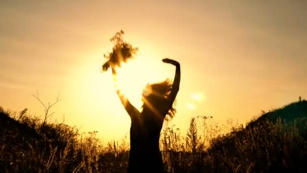 Silueta de una chica que salta alegremente y agita sus manos contra el fondo de una brillante puesta de sol con un ramo de flores silvestres en sus manos. El concepto de felicidad y libertad. 4K — Vídeo de stock