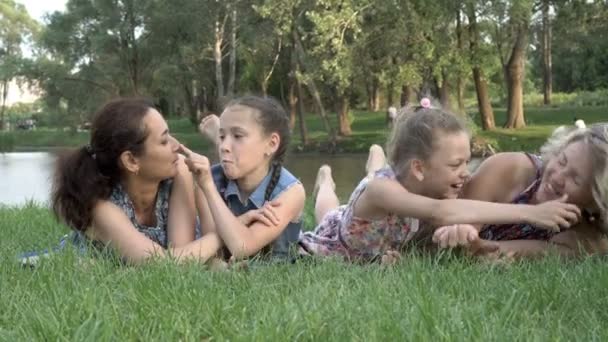 Familia del mismo sexo: madres con hijas pequeñas se divierten y juegan tumbadas en la hierba del parque al atardecer de un día de verano junto al río. Unas vacaciones en familia al aire libre. Concepto. 4K — Vídeo de stock