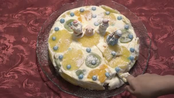 Mujer corta en pedazos pastel de Navidad con crema blanca y fruta, decorado con figuras de Santa Claus y animales. La torta vale en el plato de cristal, en los manteles rojos. Primer plano. La vista desde arriba — Vídeos de Stock