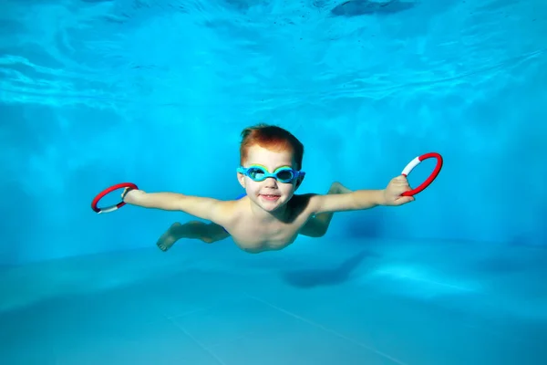 Niño deportivo nadando bajo el agua en la piscina con juguetes en sus manos. Retrato. De cerca. Fotografía submarina. Orientación horizontal — Foto de Stock