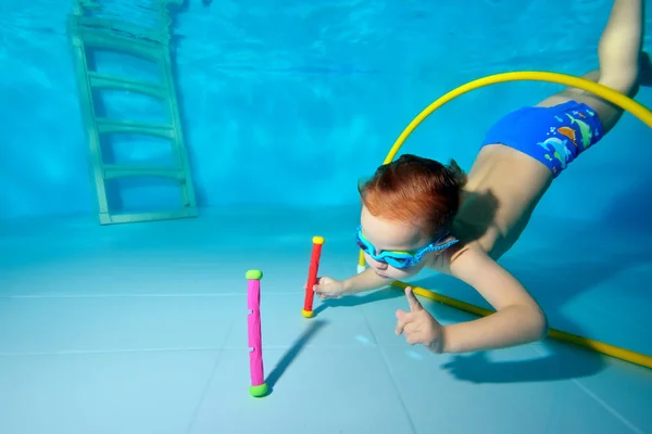 Un niño nada bajo el agua, se sumerge en el fondo de la piscina para juguetes a través de un aro amarillo. Retrato. Fotografía submarina. Orientación horizontal — Foto de Stock