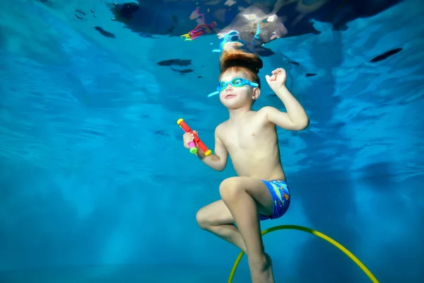 Pequeño chico deportivo nada bajo el agua, se sumerge en el fondo de la piscina para juguetes. Retrato. Foto bajo el agua. Orientación horizontal —  Fotos de Stock