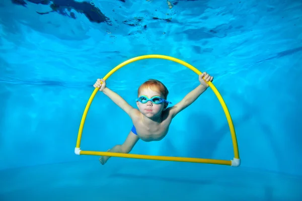 Un niño deportista se dedica a nadar en la piscina, bajo el agua en la parte inferior, posando con un aro amarillo en las manos y mirando a la cámara. Fotografía submarina. Orientación horizontal — Foto de Stock