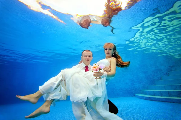 Hermosos novios bajo el agua en la piscina en un día soleado. El novio abraza y sostiene a la novia en sus brazos abajo. Retrato. Concepto. Boda bajo el agua. Vista horizontal — Foto de Stock