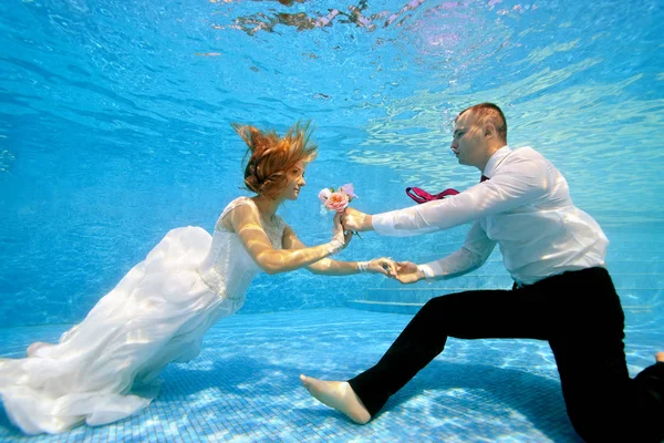 En vacker brud i en vit klänning simmar under vattnet till brudgummen och ger honom en bukett. Bruden och brudgummen belyses av solen under vattnet. Porträtt. Koncept. Undervattensfotografering — Stockfoto