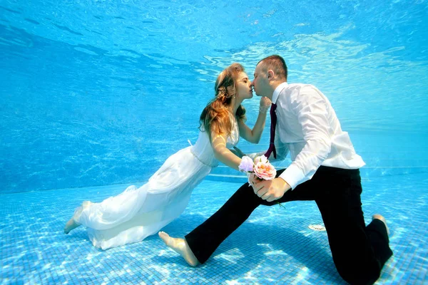 Lycklig brud och brudgum hålla händerna och kyss under vattnet på botten av poolen på en solig dag. Bröllopsbukett i handen. Porträtt. Koncept. Undervattensfotografering. Horisontell vy — Stockfoto