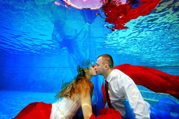 Magische Unterwasserhochzeit. glückliche Braut und Bräutigam schwimmen und küssen unter Wasser im Pool bei Sonnenuntergang, umgeben von rotem und blauem Stoff. Porträt. Konzept. Unterwasserfotografie — Stockfoto