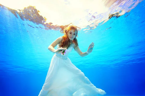 Schöne Braut mit roten Haaren schwimmt und posiert vor der Kamera unter Wasser im Pool in einem weißen Kleid mit Blumen in der Hand an einem sonnigen Tag. Porträt. Ansicht von unten. Unterwasserfotografie — Stockfoto