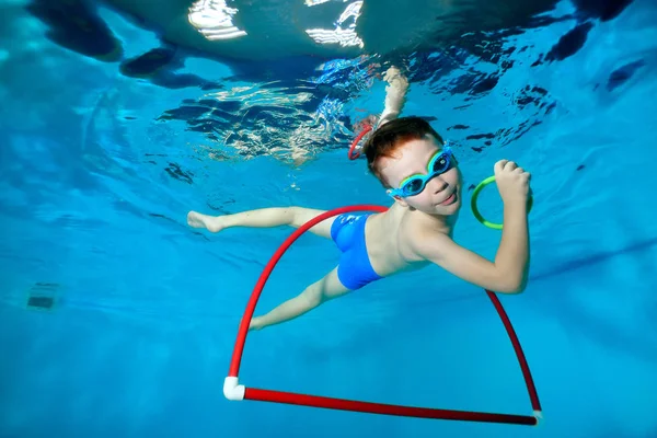 Niño deportivo, nada fuera del aro rojo bajo el agua en la piscina con juguetes en sus manos. Retrato. Fotografía submarina. Orientación horizontal — Foto de Stock