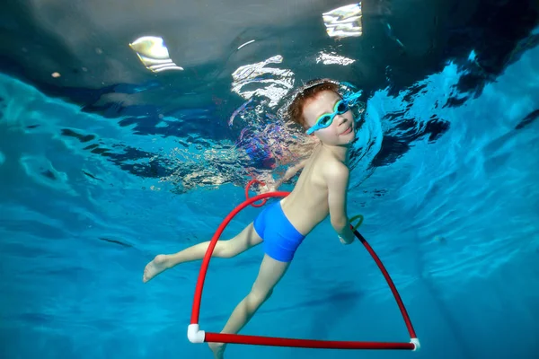 Un niño deportivo nada bajo el agua en la piscina, emerge a la superficie del aro hula y mira a la cámara. Retrato. Fotografía submarina. Orientación horizontal — Foto de Stock