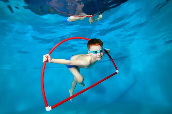Niño sonriente, nadando en la piscina, bajo el agua en el fondo, nada a través de un aro rojo con juguetes en las manos y mirando a la cámara. Fotografía submarina. Orientación horizontal — Foto de Stock
