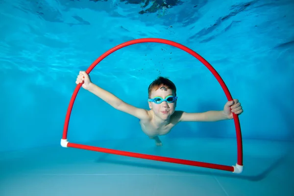 Glimlachend klein sportjongen, bezig met zwemmen in het zwembad, onderwater aan de onderkant, poseren met een rode hoepel in zijn handen en kijken naar de camera. Onderwater fotografie. Horizontale oriëntatie — Stockfoto