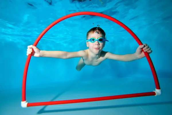 Un bambino è impegnato a nuotare in piscina, sott'acqua in fondo, posa con un cerchio rosso tra le mani e guarda la telecamera. Ritratto. Fotografia subacquea. Orientamento orizzontale — Foto Stock