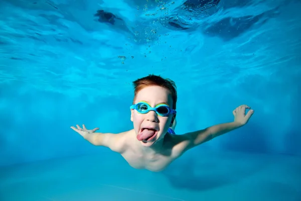 Feliz bebé sonriente, chico, nada bajo el agua en la piscina en gafas de natación y muestra la lengua en la cámara sobre fondo azul. Retrato. Fotografía submarina. Orientación horizontal — Foto de Stock