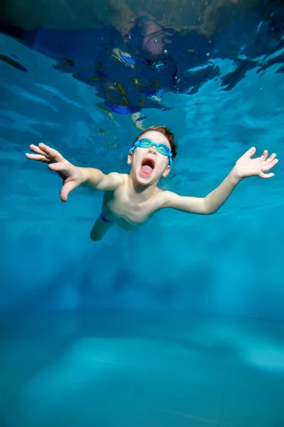 Riéndose niño deportivo nadando bajo el agua, cerca de la superficie del agua. Brazos extendidos como un pájaro. Posando para la cámara con los ojos abiertos. Encantado. Foto bajo el agua. Vista vertical — Foto de Stock