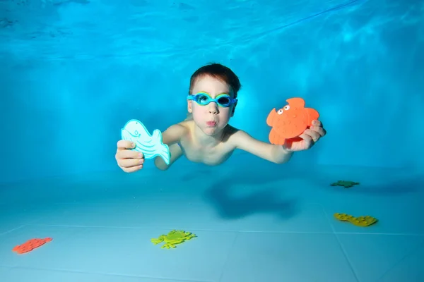 Lindo bebé deportivo nada bajo el agua y tiene juguetes marinos en su mano. Mira a la cámara y sonríe. Retrato. Fotografía submarina. Orientación horizontal — Foto de Stock