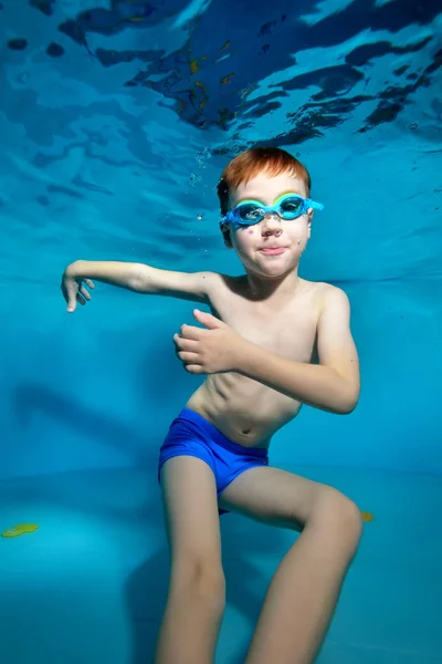 Un niño nada y juega bajo el agua en la piscina con los brazos separados y mira a la cámara. Bailando bajo el agua. Retrato. Fotografía submarina. Orientación vertical de la imagen — Foto de Stock