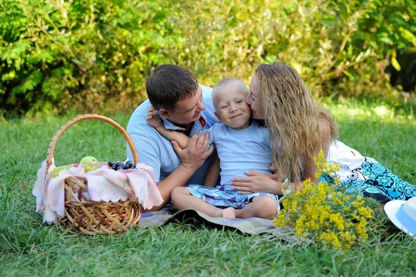 Mutlu bir aile. Küçük bir çocuk gülümsüyor ve bir yaz gününde çim üzerinde Park anne ve baba ile oturur. Oğlunu öptüğü bir anne. Doğada aile pikniği. Portre. Yatay görünüm — Stok fotoğraf