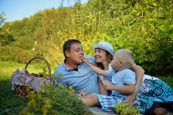 Pique-nique familial heureux dans le parc le jour de l'été. Maman, papa et un petit garçon. Le garçon nourrit les raisins du Pape et rit avec eux. Pique-nique familial dans la nature. Portrait. Vue horizontale — Photo