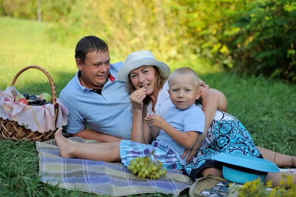 Piknik, mutlu bir aile. Anne, baba ve bebek sahilde bir sepet meyve ve çiçekler ile. Kameraya gülümsüyor. Portre. Yatay görünüm — Stok fotoğraf