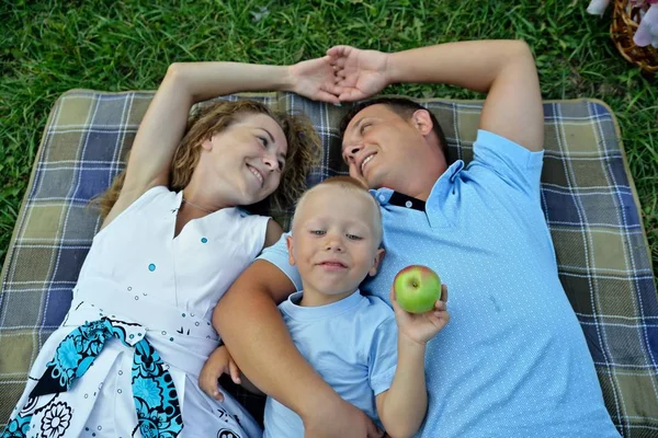 Maman, papa et petit fils se couchent et rêvent sur l'herbe dans le parc au coucher du soleil jour d'été. Les parents se regardent et sourient. L'enfant tient une pomme. Portrait. La vue du haut. Vue horizontale — Photo
