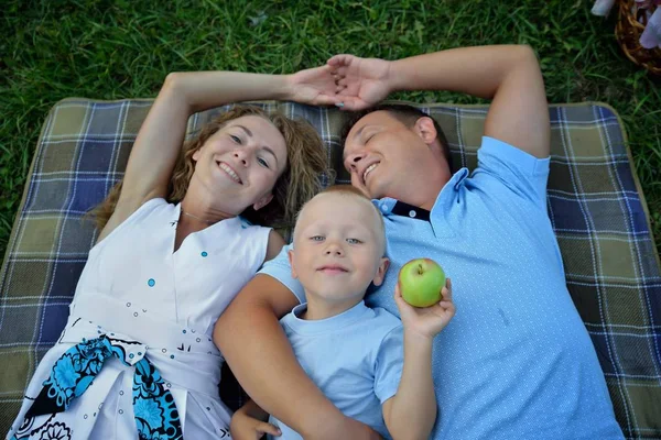 Χαρούμενη οικογένεια που ψεύδεται και ονειρεύεται στο γρασίδι στο πάρκο την καλοκαιρινή μέρα του ηλιοβασιλέματος. Οι γονείς κρατούν τα χέρια και χαμογελούν. Το παιδί κρατά ένα μήλο. Πορτρέτο. Η θέα από την κορυφή — Φωτογραφία Αρχείου
