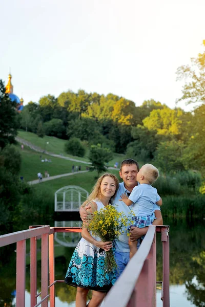 Bonne famille : père, mère et enfant se tiennent dans le parc au bord de l'eau au coucher du soleil de la journée d'été. Le garçon embrasse le Pape. La famille se promène au grand air. Portrait. Vue verticale — Photo