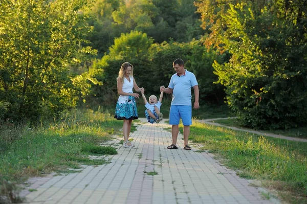 Les parents balancent le petit garçon dans leurs bras comme une balançoire et marchent le long du chemin dans le parc au coucher du soleil d'une journée d'été. Le concept du bonheur familial. Tais toi. Vue horizontale — Photo