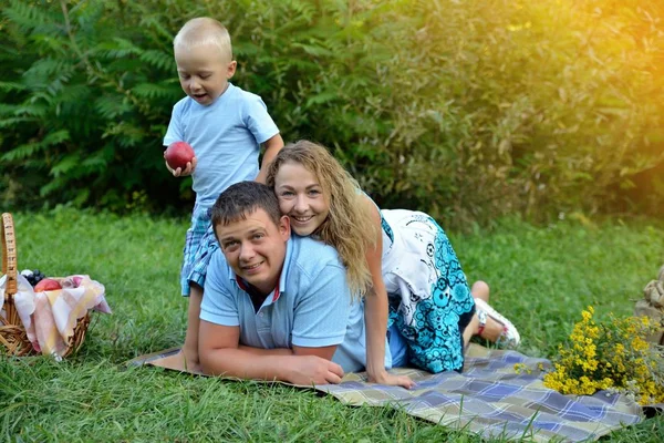 新鮮な空気の中で家族のピクニック。お父さん、お母さんは毛布の上に横たわ、小さな息子がアップルの隣に立っている。日没時に公園で休む。家族の幸福の概念。肖像 画。水平方向のビュー — ストック写真