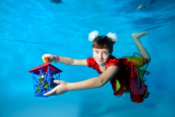 Una linda niña con grandes ojos nada bajo el agua en la parte inferior de la piscina en un vestido rojo y muestra el juguete a la cámara. Retrato. Vista horizontal — Foto de Stock