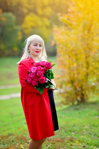 Cute middle-aged woman, blonde standing in the Park in a red coat with a bouquet of scarlet roses on the background of autumn trees. He looks away thoughtfully. Size plus xxl female. Vertical view