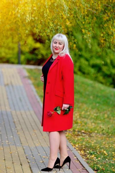 Una hermosa rubia grande está de costado en el camino en el Parque con un vestido negro y un abrigo rojo con una rosa en las manos. Talla de mujer más xxl. Plan general. Vista vertical —  Fotos de Stock