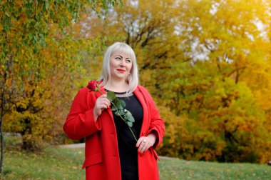 Kırmızı bir ceket büyük bir güzel sarışın ve bir kızıl gül elinde, parkta bir güneşli sonbahar günü yan rüya görünüyor. Kadın büyüklüğü artı XXL. Yatay görünüm